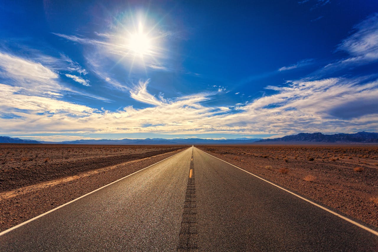 A long, straight road stretches across a dry desert landscape under a bright blue sky and shining sun.